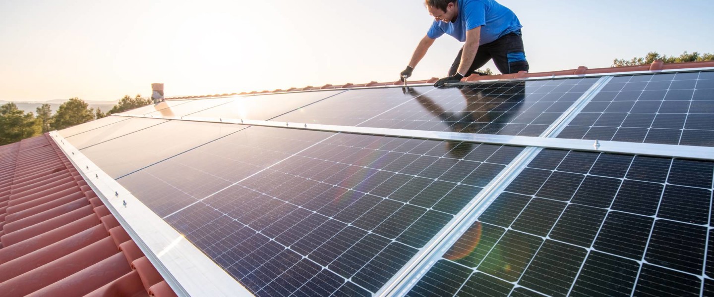 A person installing solar panels on a sloped red tiled roof, with the sun shining in the background.