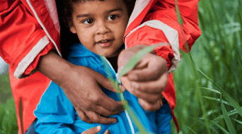 An adult in a red jacket holds a child who is holding a blade of grass, surrounded by green fields.