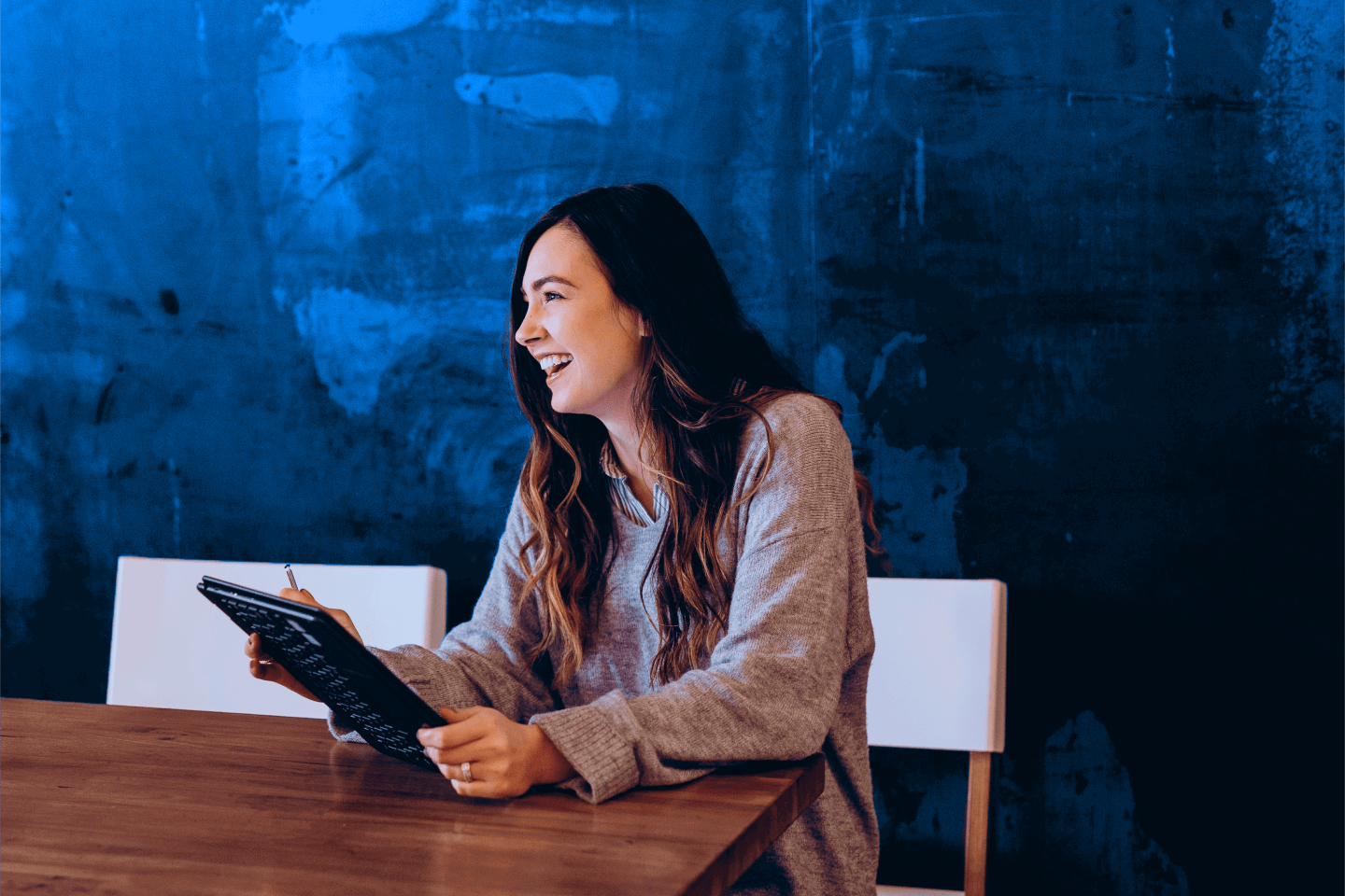 A woman in a gray sweater sits at a wooden table, looking at a tablet, with a blue wall in the background.