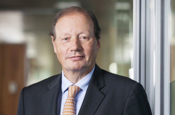 Mr D.M. Sluimers in a black suit with an orange tie, standing in front of a modern office wall, with a blurred background.