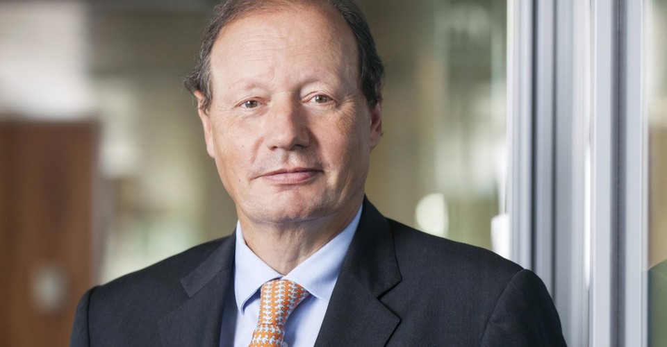 Mr D.M. Sluimers in a black suit with an orange tie, standing in front of a modern office wall, with a blurred background.
