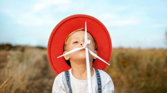 A child in a red hat stands in a field of yellow grass, looking playful with an arrow in its mouth.