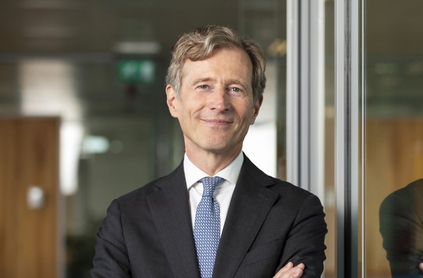Jeroen in a black suit and blue tie stands with his arms crossed in front of a glass wall in an office setting.