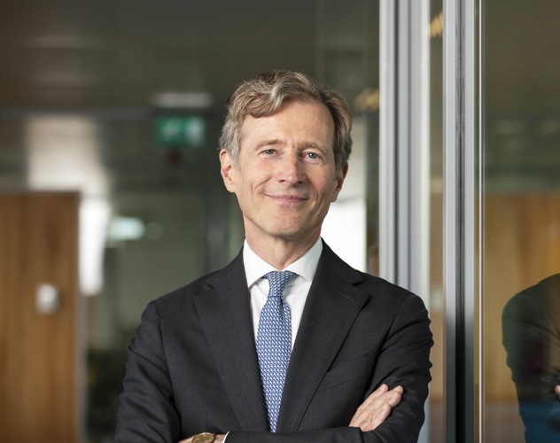 Jeroen in a black suit and blue tie stands with his arms crossed in front of a glass wall in an office setting.