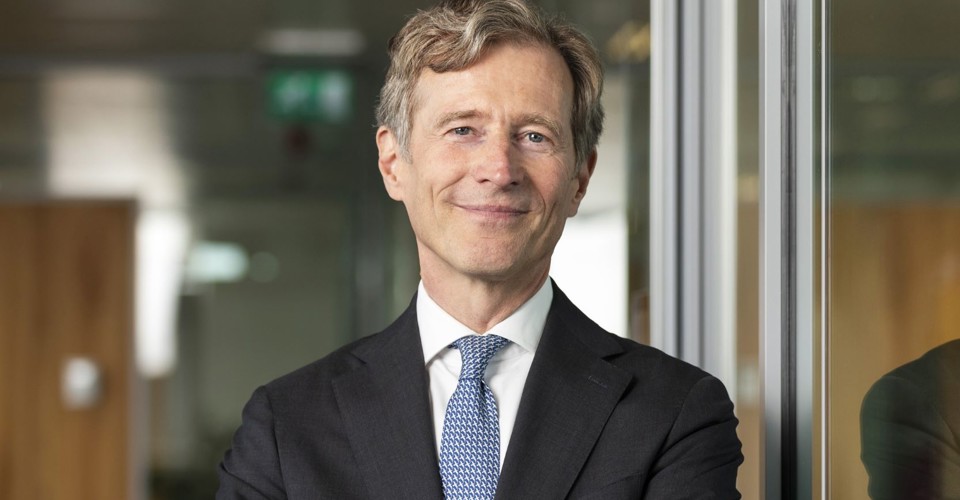 Jeroen in a black suit and blue tie stands with his arms crossed in front of a glass wall in an office setting.