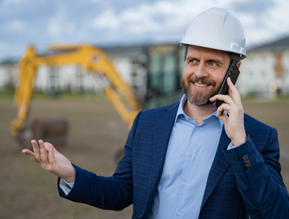 A man in a blazer stands on a construction site, speaking on his phone, with an excavator visible in the background.