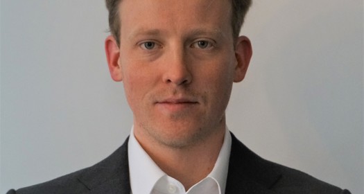 A man in formal attire wearing a black blazer and a white shirt, positioned against a neutral background.