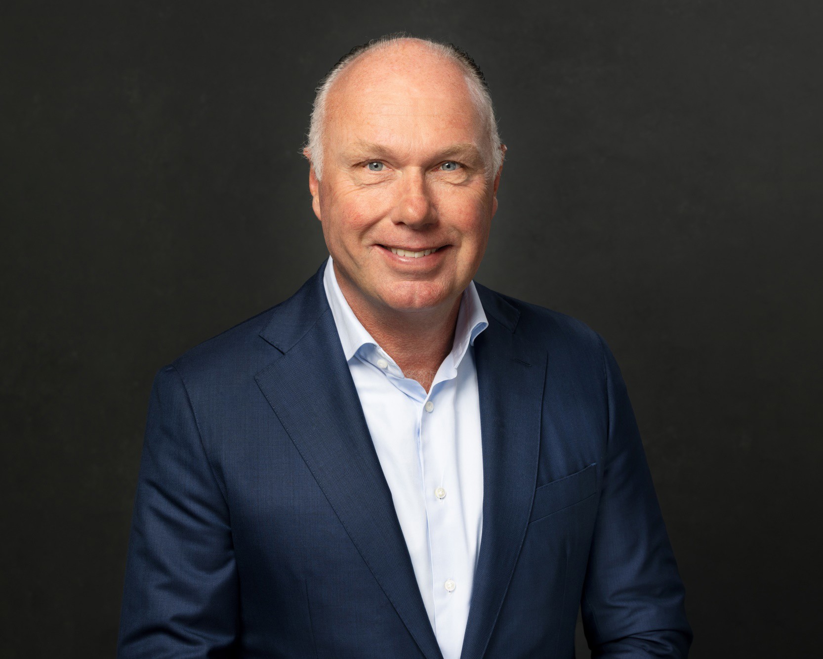 Nick in a dark blue suit with a light blue shirt stands against a black background.