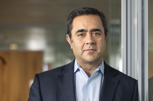 Qasim in a dark blazer and light blue shirt stands next to a glass wall in a modern office setting.
